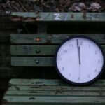 Close-up of an analog clock on a rustic green wooden bench outdoors.